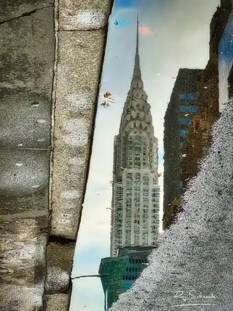 A puddle on a city sidewalk reflects the Chrysler Building, with its iconic spire and ornate facade clearly visible in the…