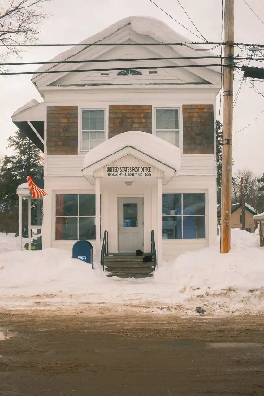 A snowy, two-story white post office building in Constableville, New York, with snow piled on its roof and steps. Two black…