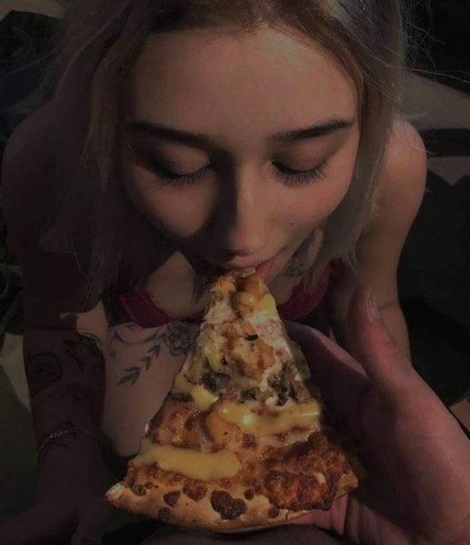 A person with light-colored hair and visible tattoos on their arm is holding a large slice of cheese pizza with both hands…
