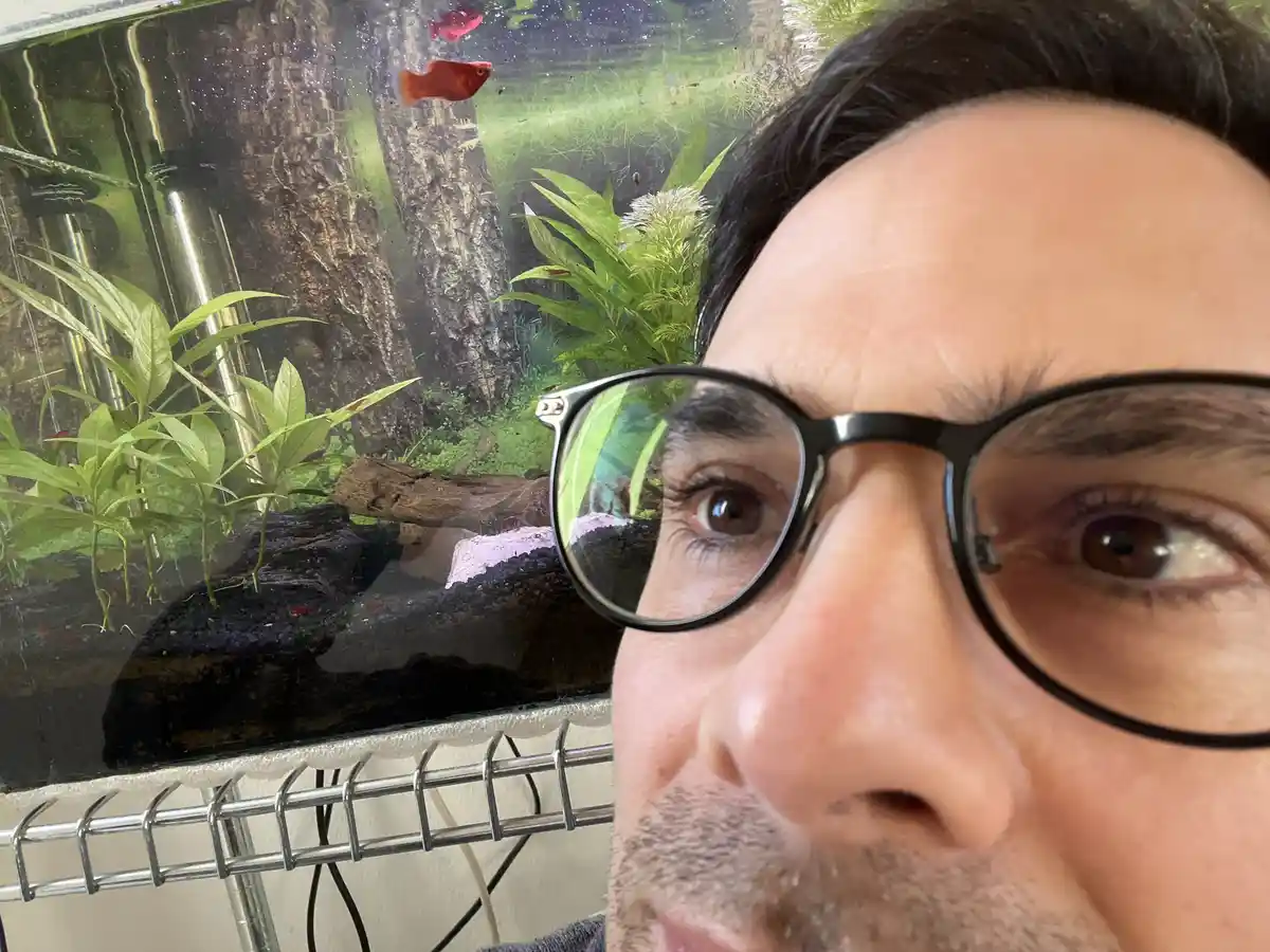 A close-up selfie of a man with dark hair and black-rimmed glasses, looking directly at the camera with a neutral…