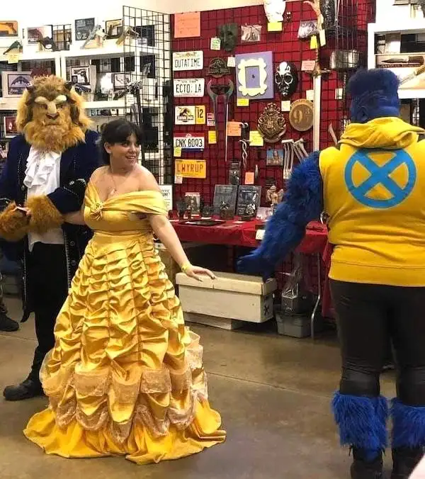 A woman in a yellow Belle costume from Beauty and the Beast stands between a man in a Beast costume and another man in a…