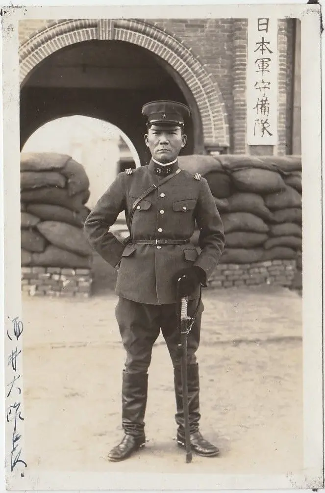 A vintage black-and-white photograph shows a stern-looking Japanese soldier in uniform standing at attention with hands on…
