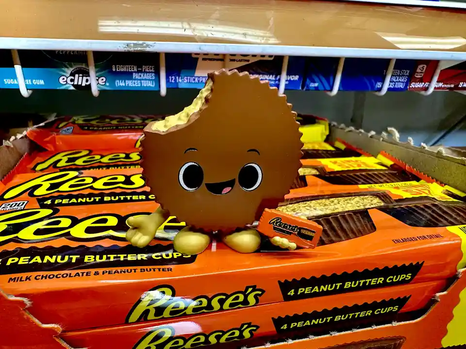 A cheerful, cartoonish Reese's Peanut Butter Cup character with large eyes and a smile sits atop a display of Reese's Peanut…