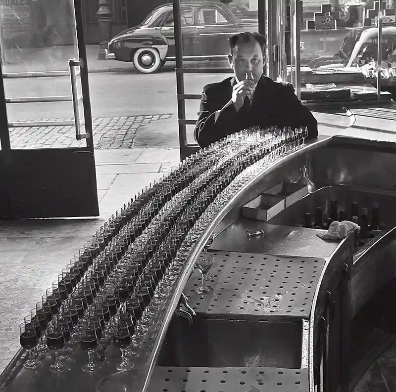 A black-and-white photo shows a man in a suit sitting behind a curved bar, sipping from a glass while staring intently. The…