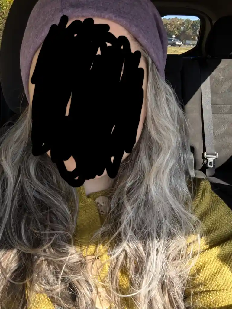 A person with long, wavy, platinum blonde hair sits in a car wearing a purple beanie and a yellow sweater, their face…