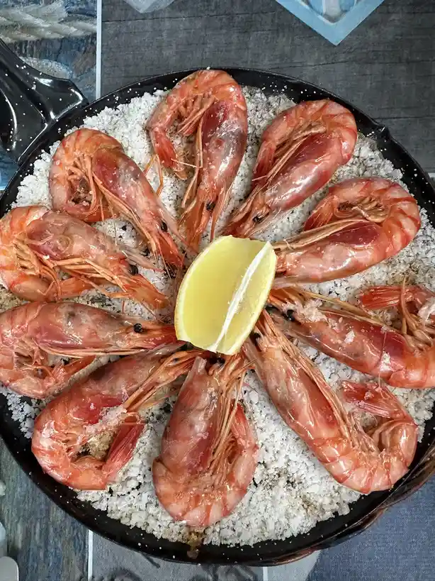 A black skillet contains a ring of cooked, pink shrimp arranged around a central wedge of lemon, all resting on a bed of…