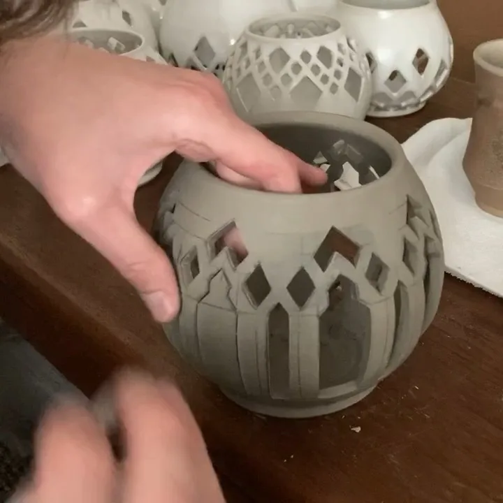 A person's hand is shown holding a gray, intricately carved clay pot with geometric cutouts, while another hand is blurred…