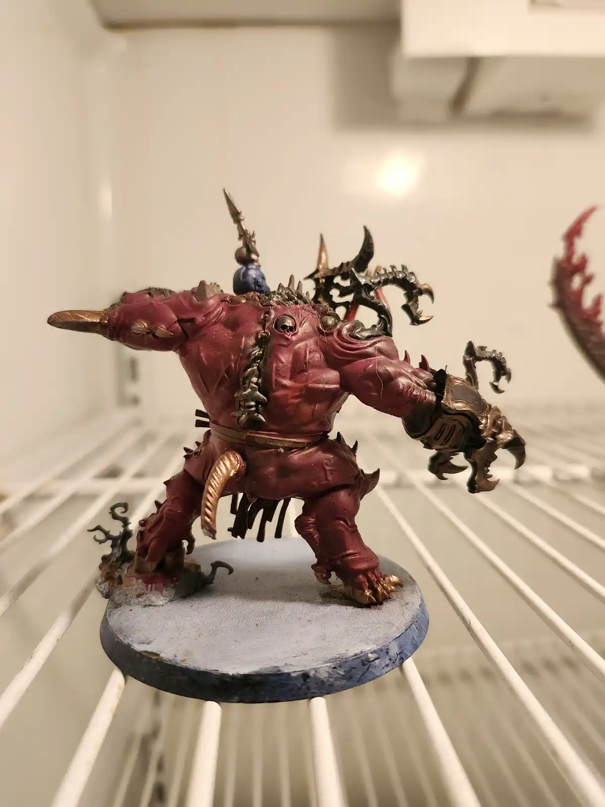 A heavily painted, muscular red fantasy monster miniature with spikes, claws, and a skull on its back stands on a blue base…