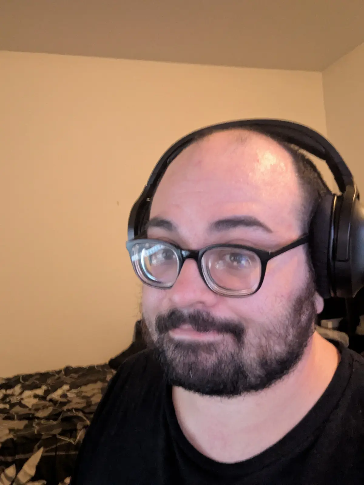A man with glasses and a beard wearing large black headphones looks directly at the camera with a slightly confused or…