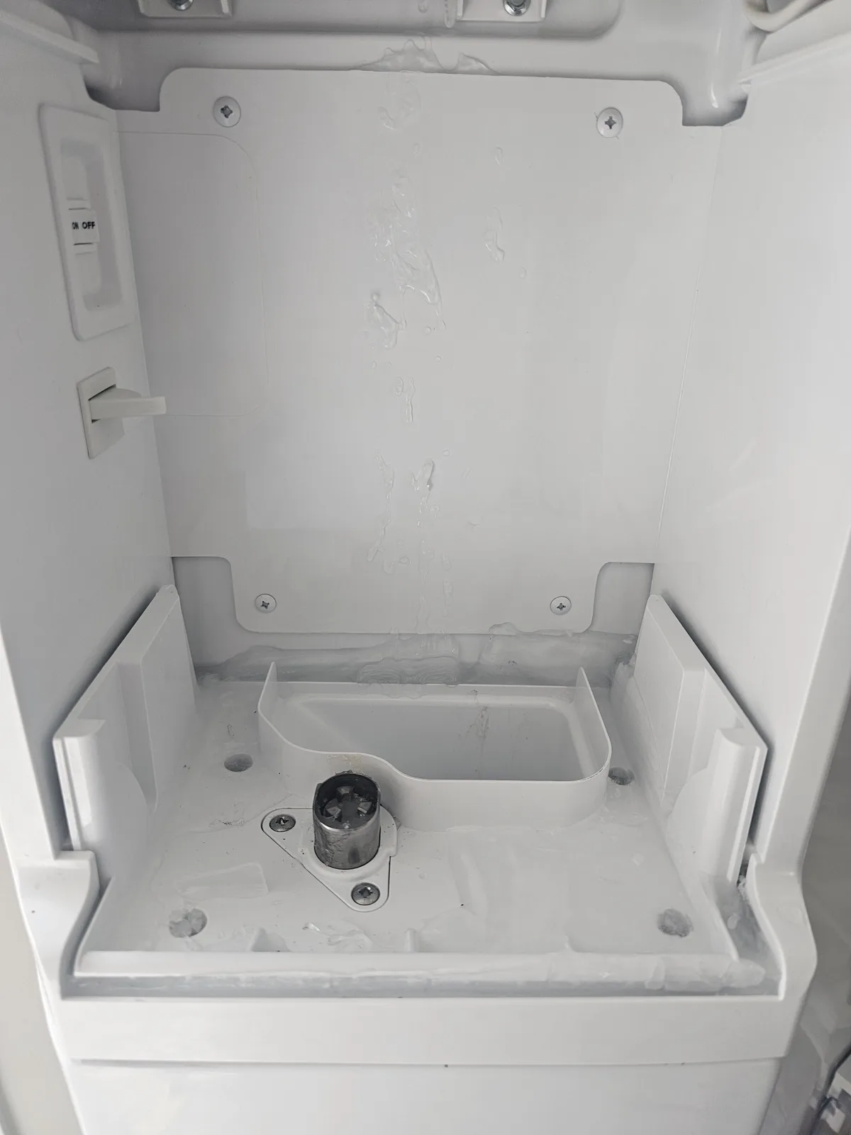 The image shows the interior of a white ice maker unit with water droplets and ice residue on the walls and tray. A metal…