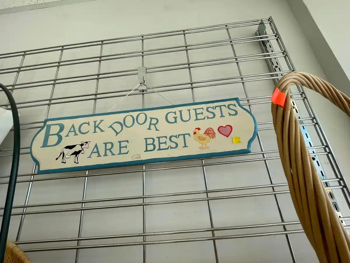 A rustic wooden sign with blue lettering and farm animal illustrations hangs on a metal grid shelf in a store. The sign…