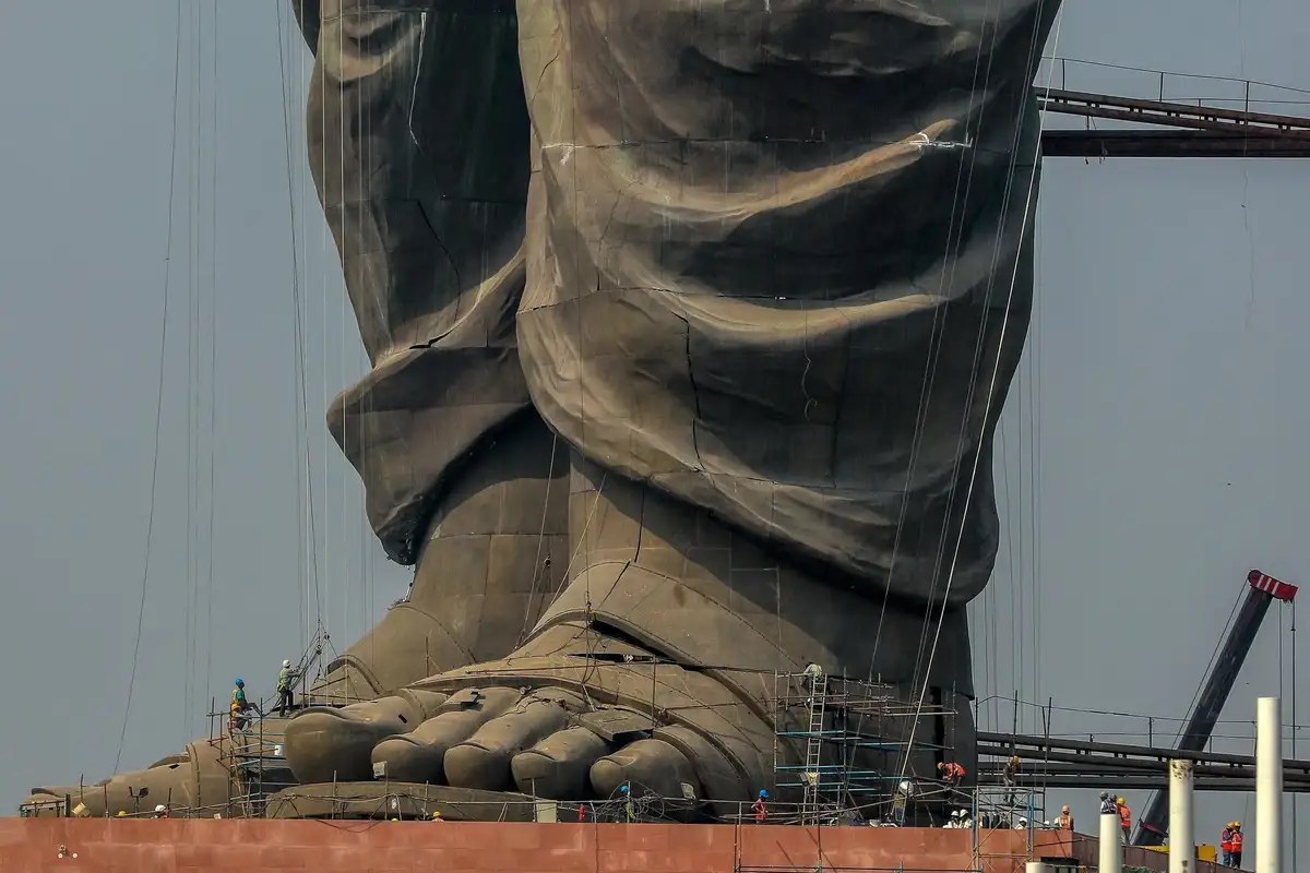 A close-up of the massive, unfinished feet of a giant statue under construction, covered in dark protective sheeting and…