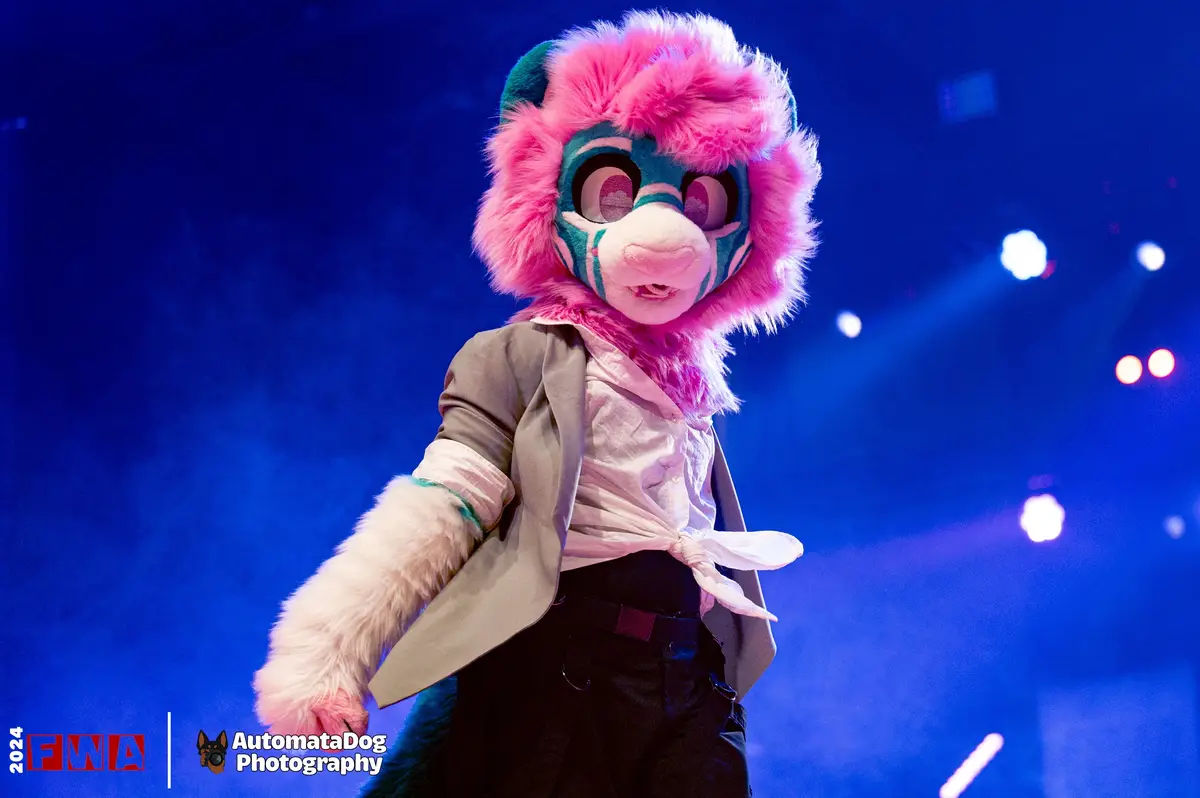 A person in a colorful furry costume with pink and teal fur, large eyes, and a fluffy mane stands on a stage under blue…