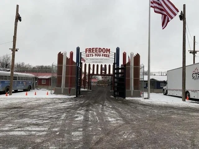 A snowy, overcast outdoor scene shows a large, decorative metal gate with the words 'FREEDOM SETS YOU FREE' in red letters…