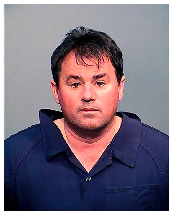 A mugshot-style photo of a man with dark hair and a neutral expression, wearing a dark blue collared shirt against a plain…