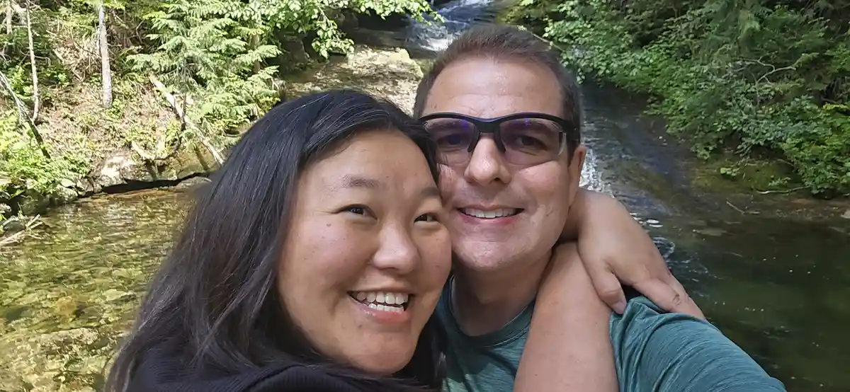A selfie of a smiling man and woman embracing near a stream with a small waterfall in the background, surrounded by lush…
