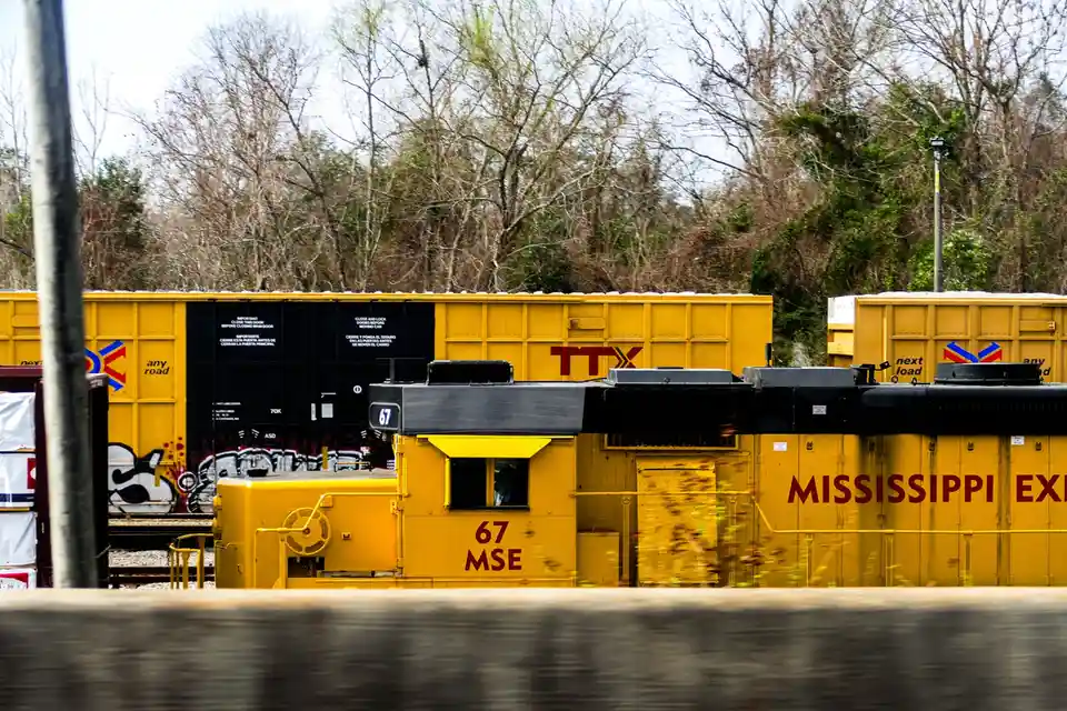 A yellow Mississippi EX train car, marked with the number 67, is captured in motion blur as it passes by. Behind it, another…