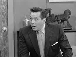 A black-and-white still of a man in a suit with a shocked expression, mouth open and eyes wide. Behind him, a doorway is…