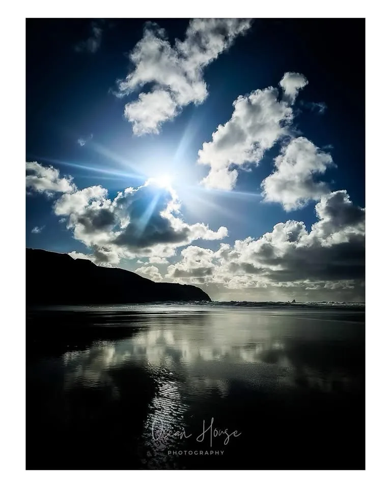 A dramatic beach photograph shows a bright sun shining through scattered white clouds against a deep blue sky, with the…