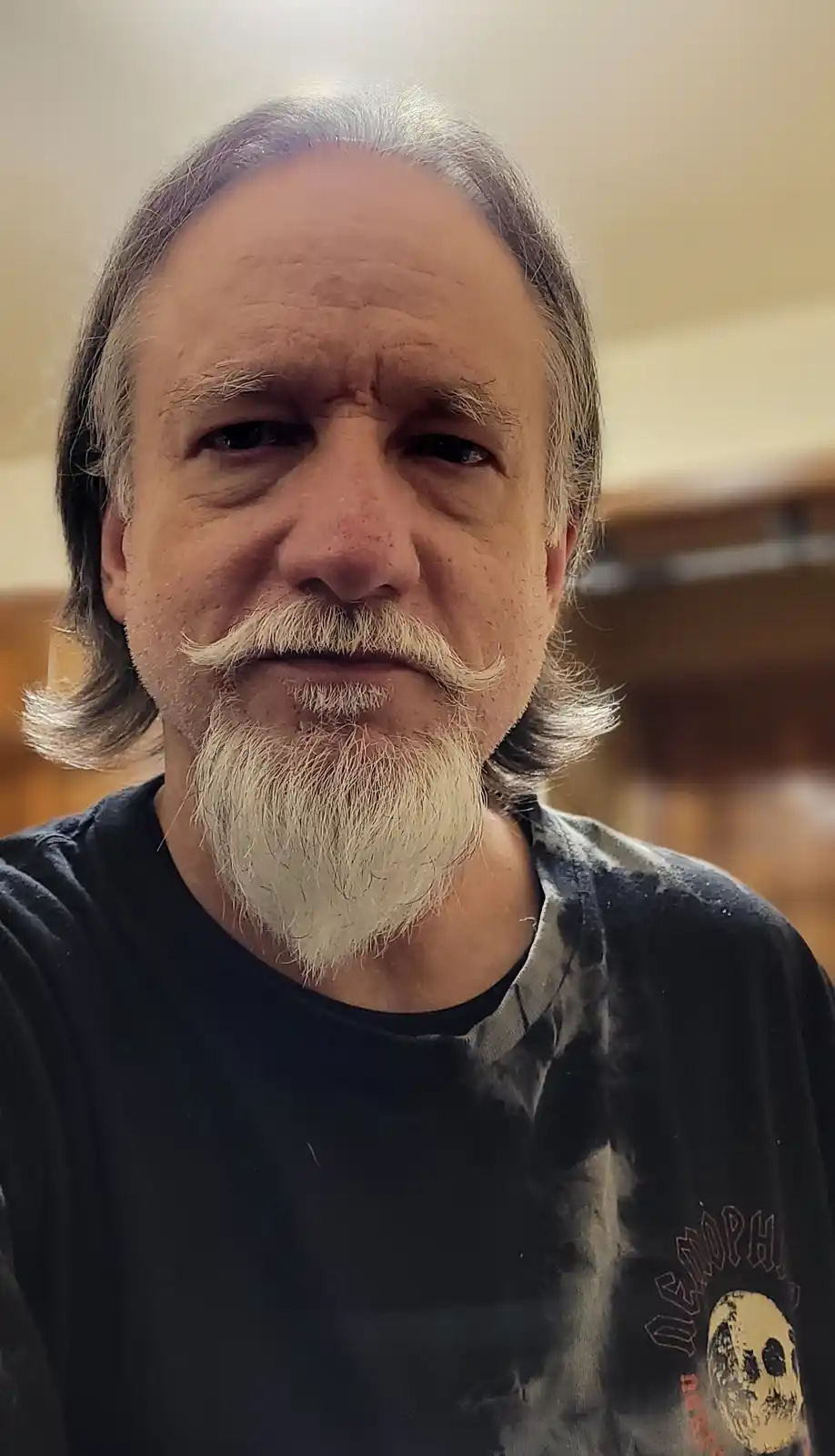 A close-up photo of a middle-aged man with long gray hair, a white goatee, and a distinctive handlebar mustache. He wears a…