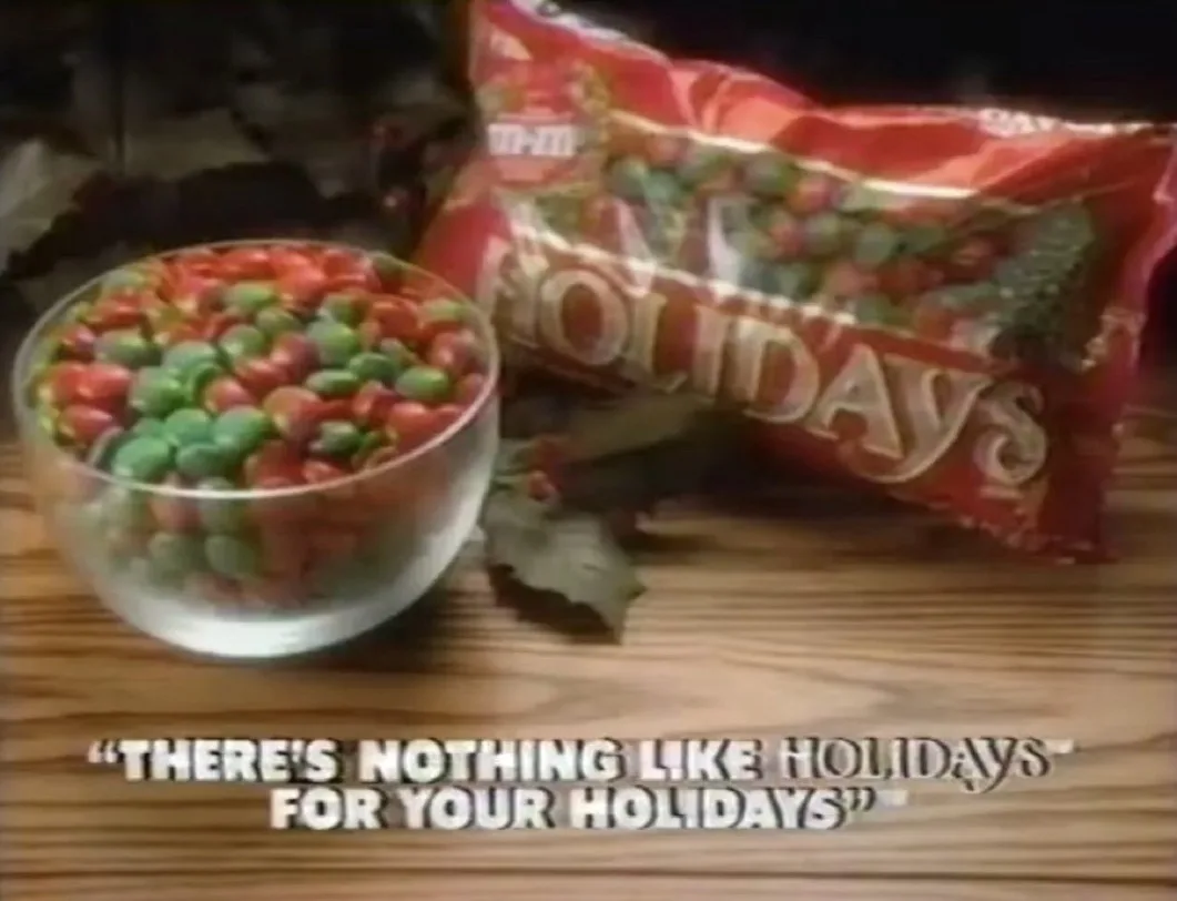 A vintage M&M's Holidays commercial still shows a glass bowl and a red bag filled with red and green holiday-themed M&M's…