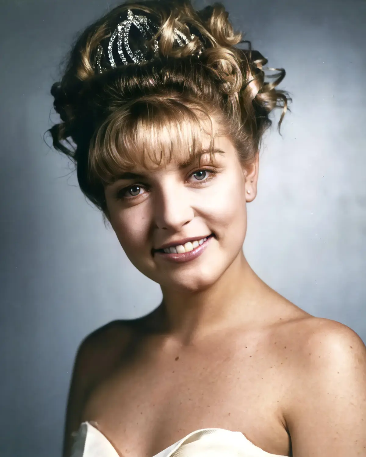 A young woman with blonde hair styled in curls and wearing a sparkly tiara smiles at the camera. She is wearing a strapless…