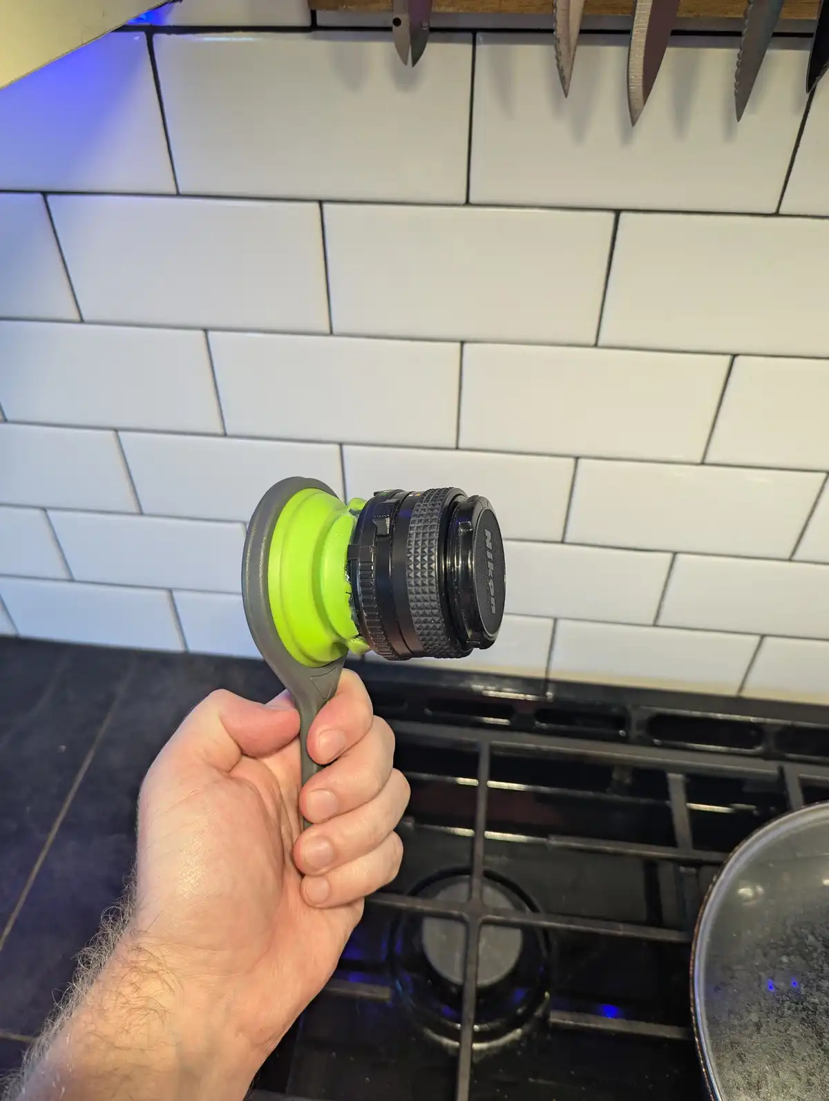 A hand holds a bright green silicone spatula with a black Nikon camera lens attached to its head, set against a kitchen…