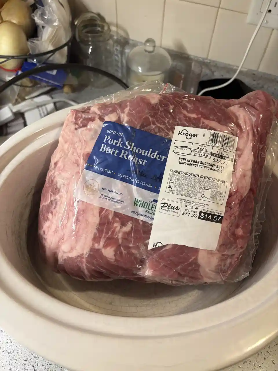 A large, bone-in pork shoulder butt roast in its plastic packaging sits inside a white ceramic slow cooker on a kitchen…