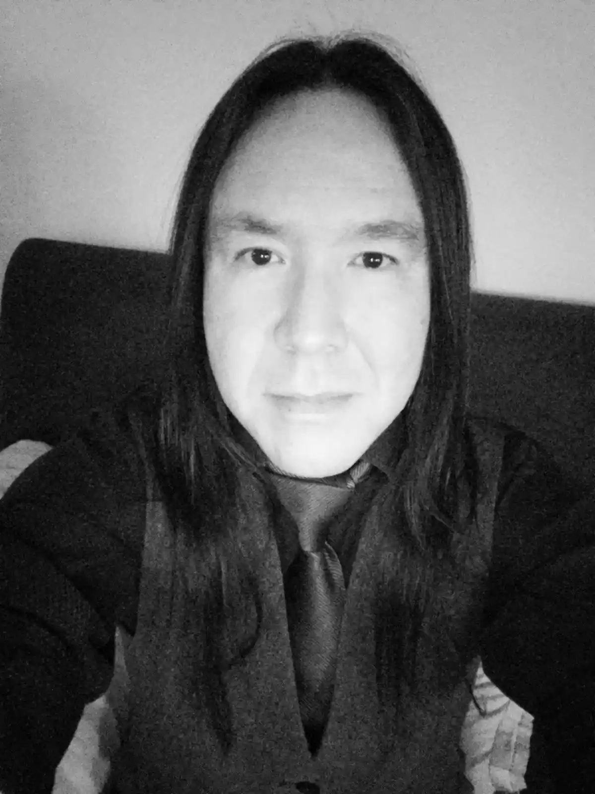 A black-and-white selfie of a person with long dark hair, wearing a dark vest and tie, seated in a chair. The image has a…
