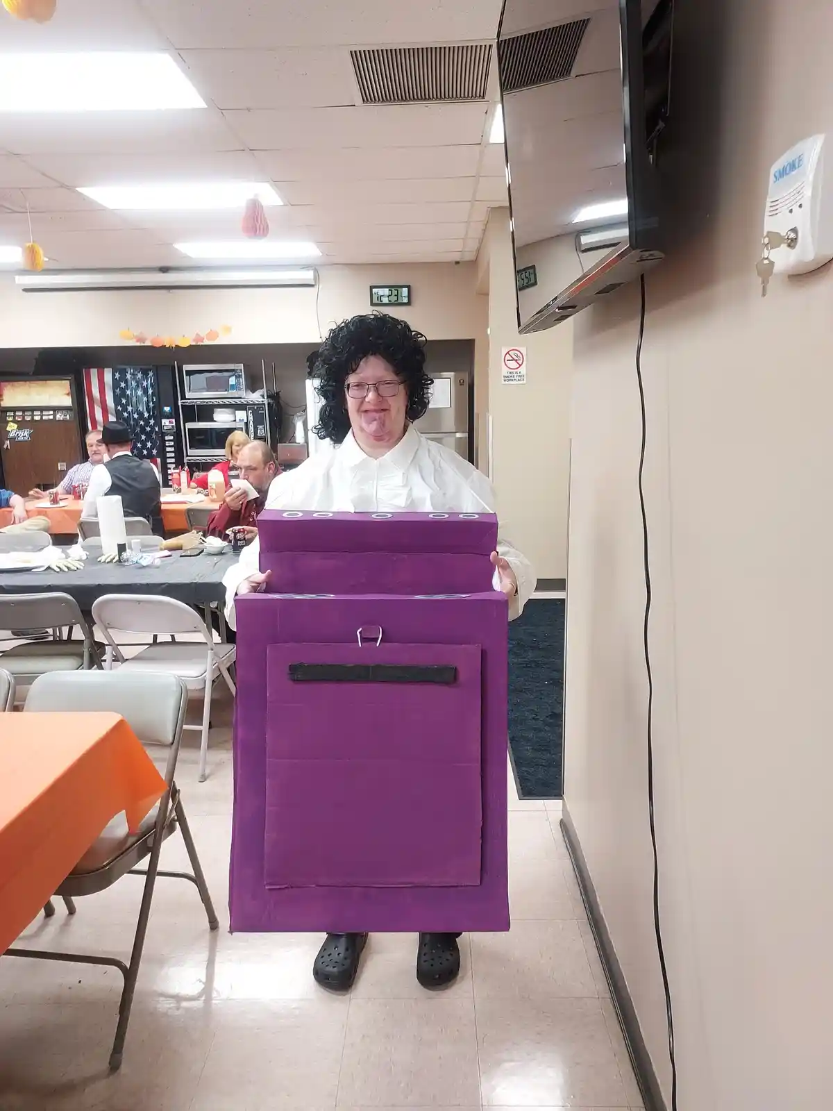 A person wearing a black wig, glasses, and a white shirt stands in a room with tables and chairs, holding a large purple…