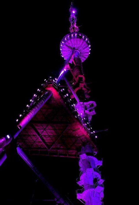 A low-angle nighttime photo of a tall, illuminated tower structure, possibly a communications or observation tower, bathed…