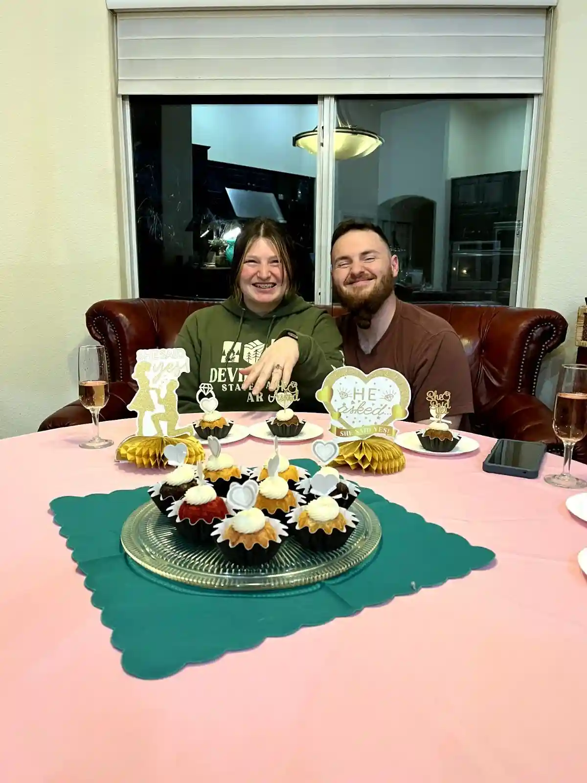 A smiling couple sits at a pink-clothed table decorated with engagement-themed cupcakes and toppers. The woman wears a green…
