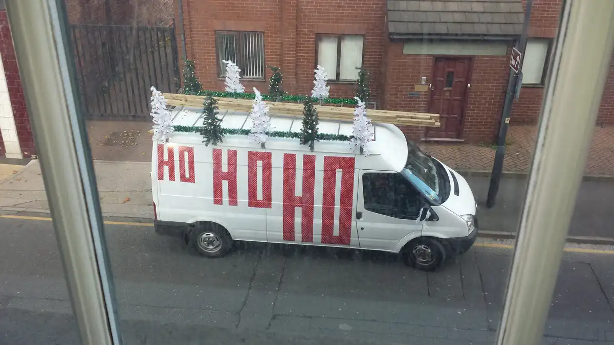 A white van parked on a street is decorated with Christmas trees and garlands on its roof and sides, with the phrase 'HO HO…