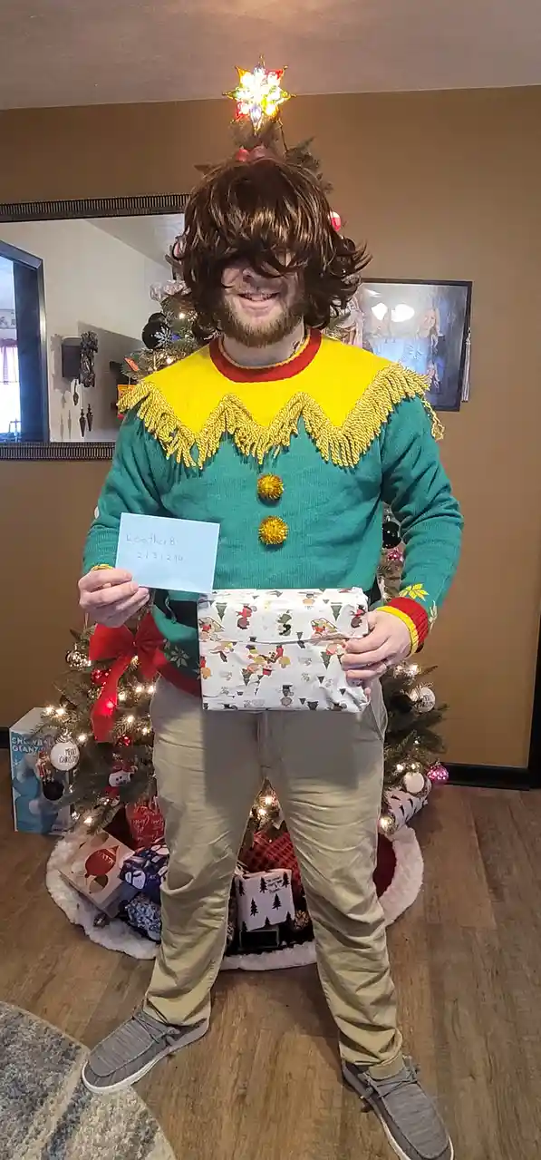 A man wearing a festive green and yellow elf sweater with a Christmas tree top hat stands in front of a decorated tree…