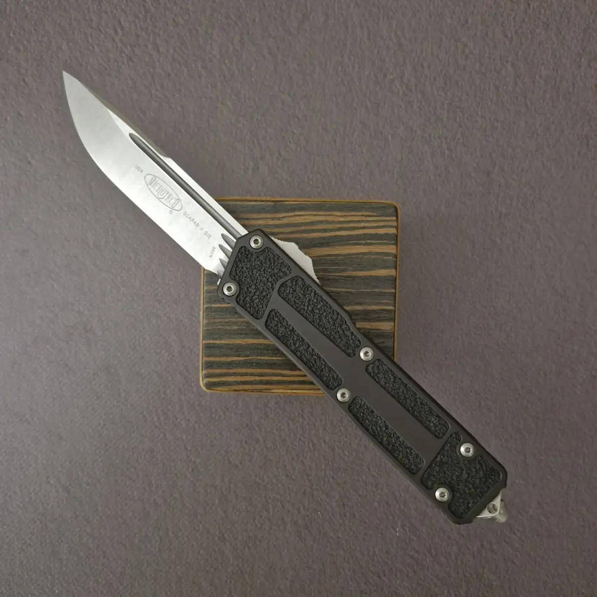 A silver-bladed Microtech folding knife with a black textured handle rests diagonally on a wooden block against a gray…