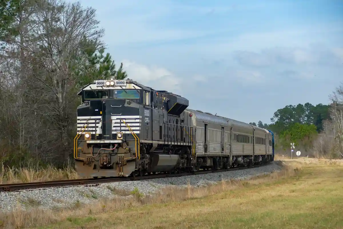 A black and white striped CPR locomotive numbered 9000 pulls a silver passenger train through a rural landscape with trees…