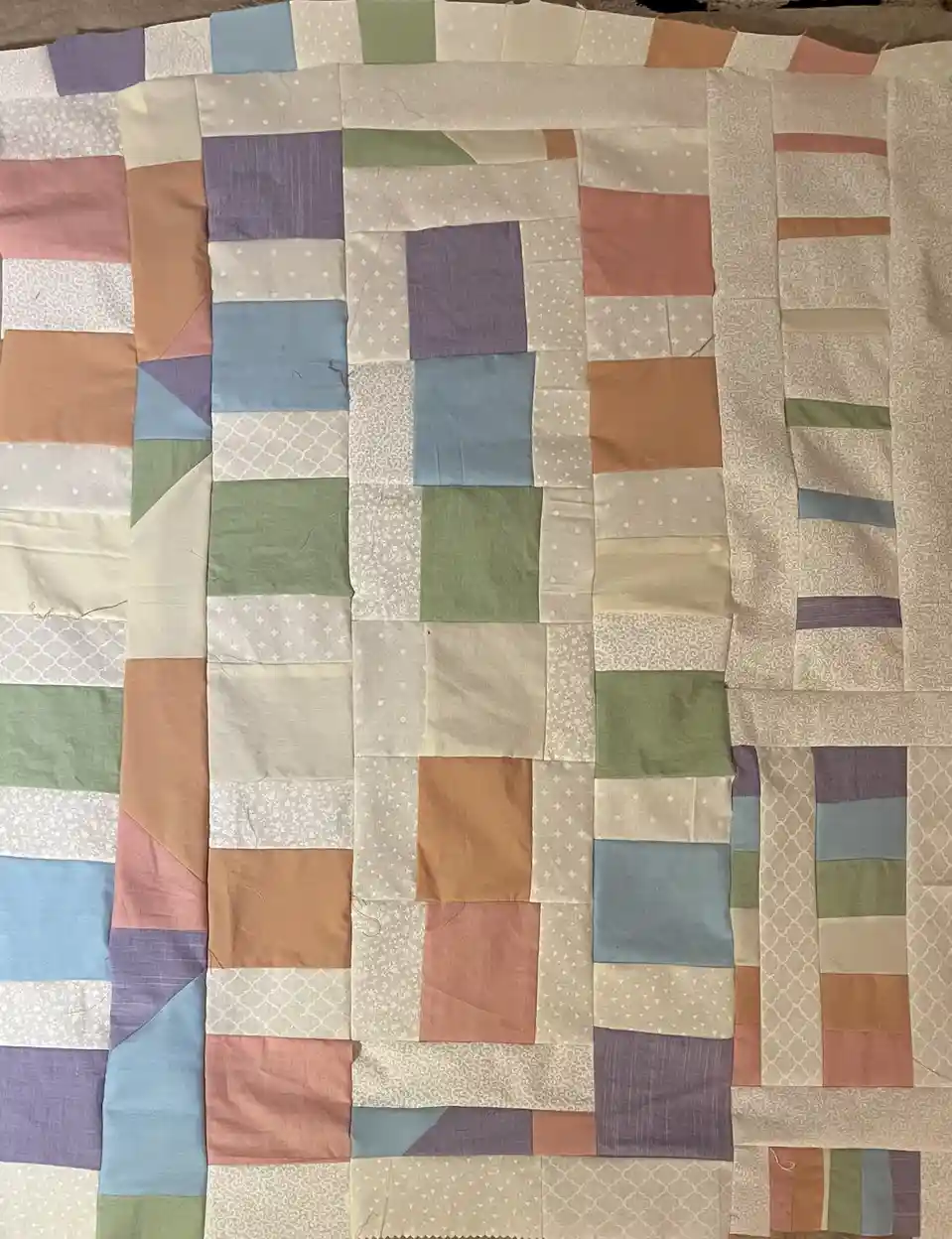 A top-down photo of a handmade patchwork quilt made of many small, irregularly shaped fabric squares in muted colors like…