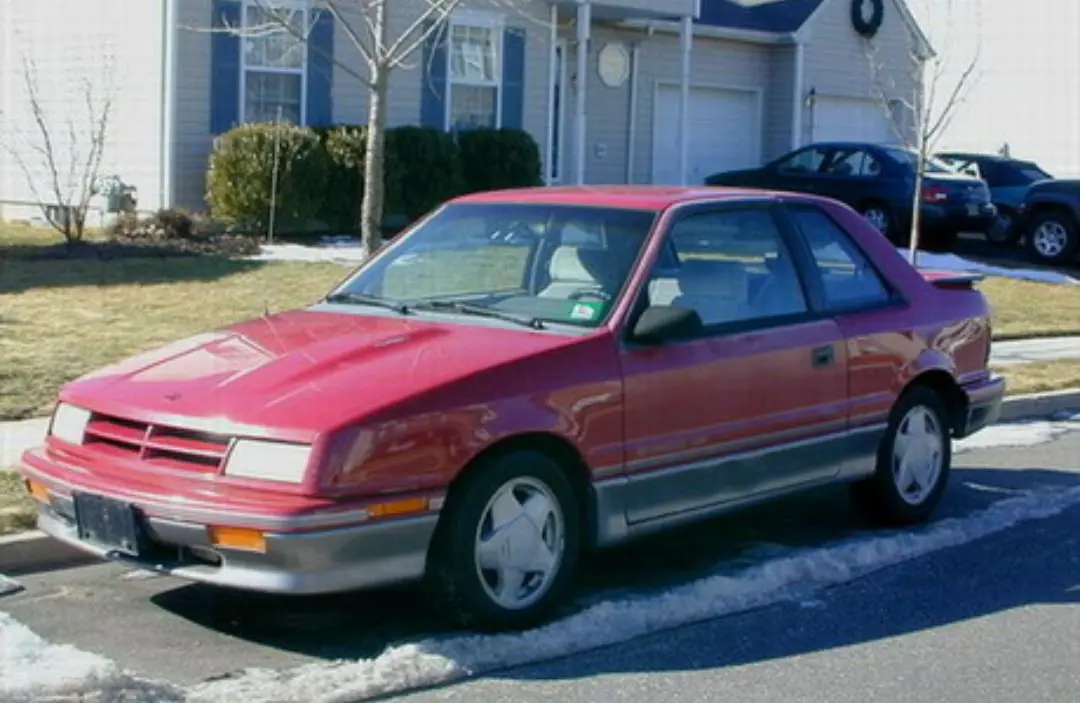 A red and silver 1980s-style two-door coupe is parked on a snowy residential street in front of a house. The car has a…