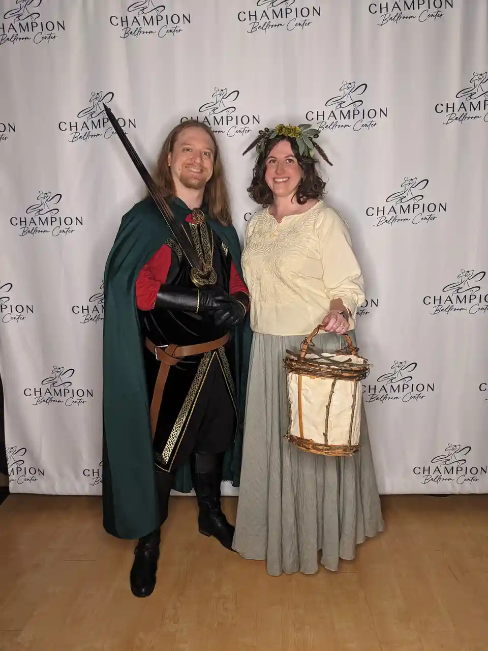 Two people dressed in medieval-style costumes pose in front of a 'Champion Ballroom Center' backdrop. The man on the left…