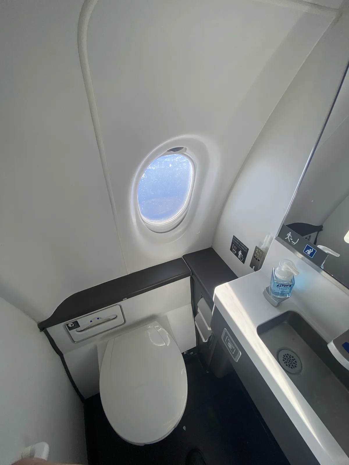 A photo of a cramped airplane lavatory showing a white toilet, a small sink with a Purell soap dispenser, and an oval…