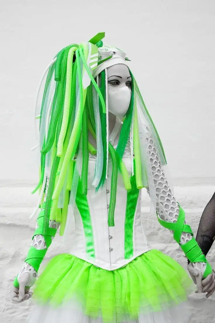 A person in a white and neon green costume with a mask and green ribbons for hair stands against a white background. The…