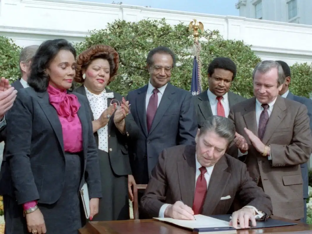 A historical photo shows President Ronald Reagan signing a document outdoors, surrounded by several smiling and applauding…