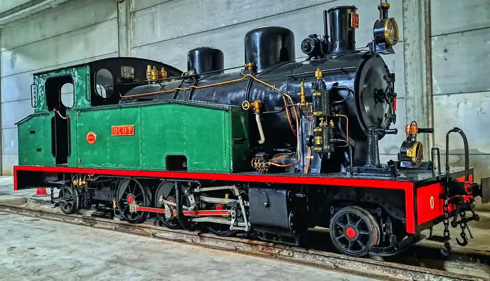 A vintage green and black steam locomotive with red trim and brass fittings is displayed indoors on tracks, with the word…