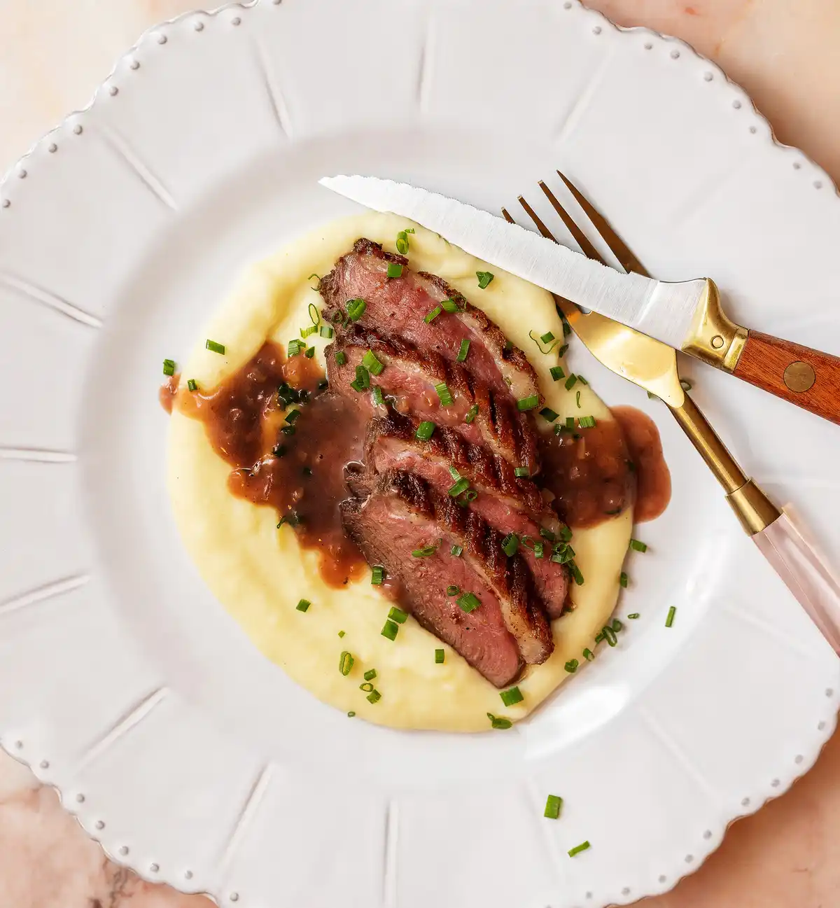 A top-down photo of a plated meal featuring sliced medium-rare meat over creamy mashed potatoes, garnished with chopped…
