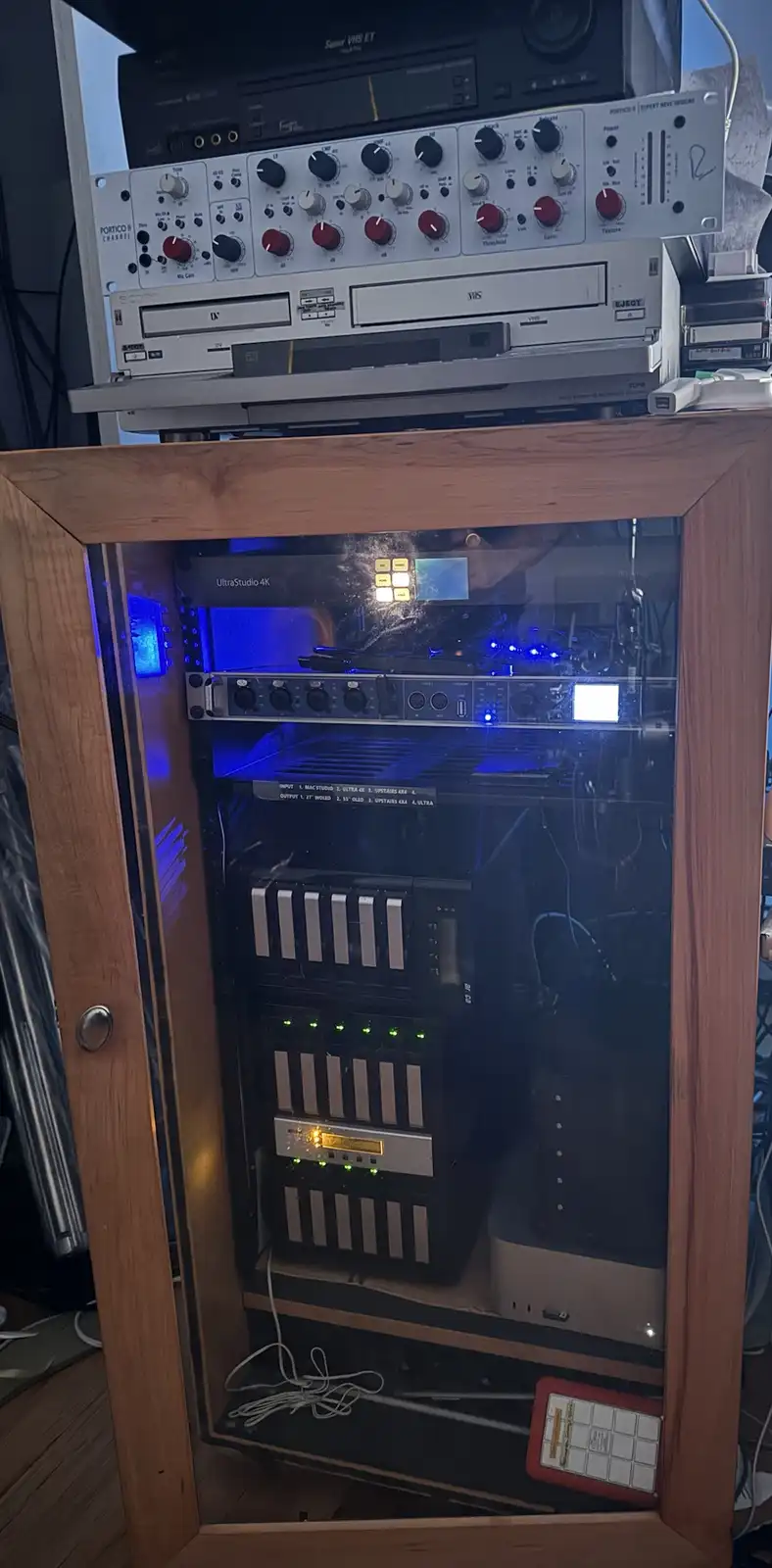A wooden cabinet with glass doors reveals a home studio setup with glowing blue lights, including an 'UltraStudio 4K' device…