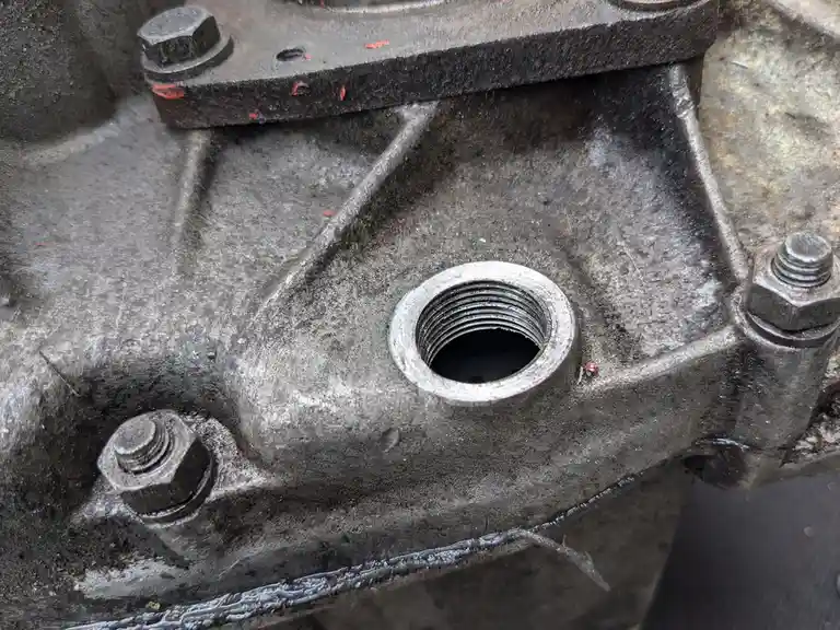 A close-up photo of a dirty, greasy metal engine block with a threaded bolt hole and three hex bolts. The overlaid text…