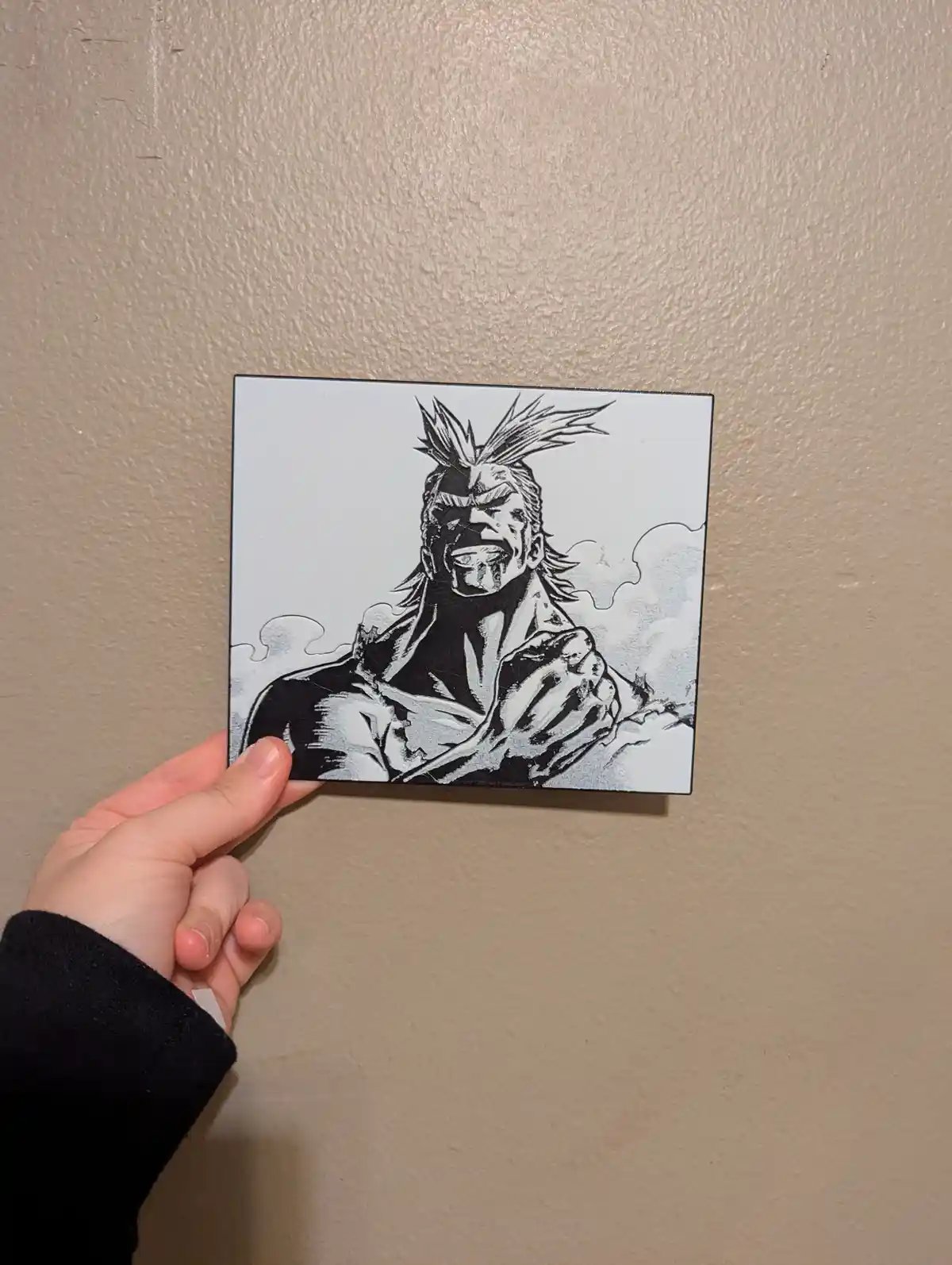 A hand holds a square black-and-white illustration of a muscular anime character with spiky hair, gritted teeth, and a…
