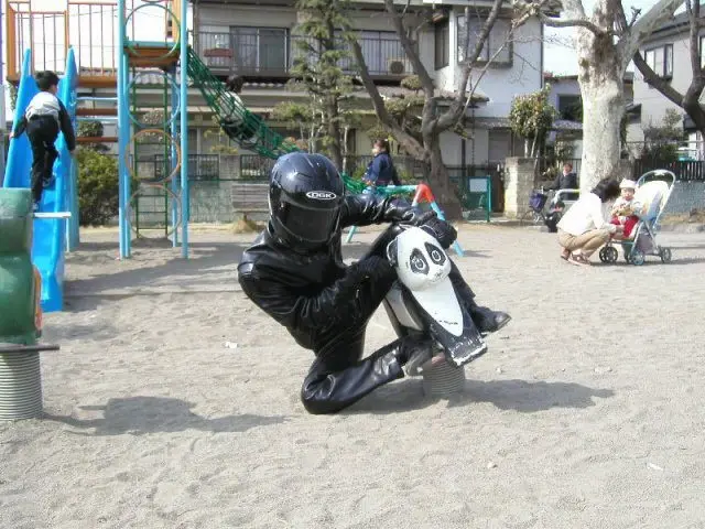 A person in full black motorcycle gear, including helmet and leather suit, is kneeling on a small white toy motorcycle in a…