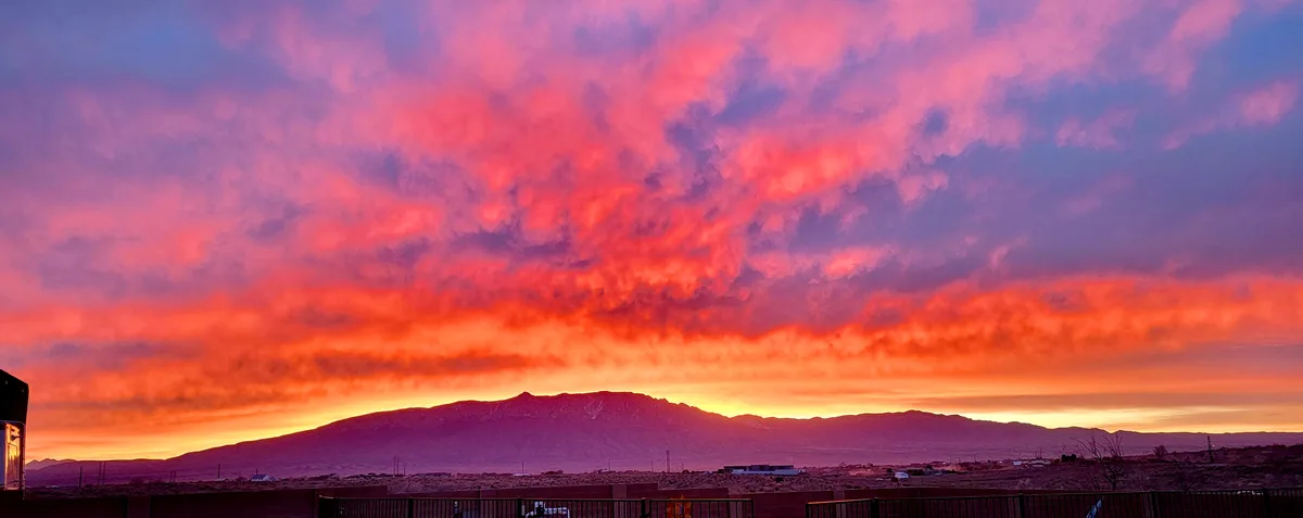 A panoramic photo of a dramatic sunset over a mountain range, with clouds glowing in fiery reds, oranges, and purples. The…