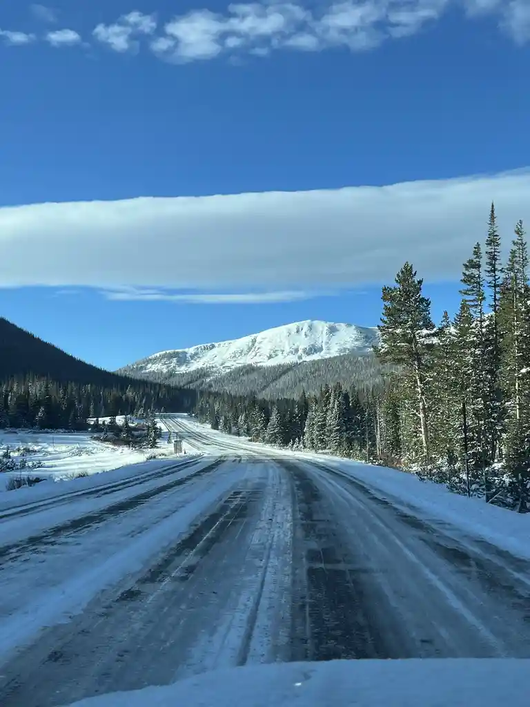A snowy mountain road stretches toward a snow-capped peak under a blue sky with a long, flat cloud. Evergreen trees line the…